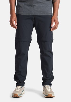 Pantalons convertibles noirs en tissu léger, avec des cuisses zippées et des poignets élastiques. Associés à des baskets grises et un t-shirt gris foncé.