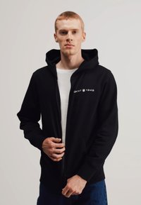 Felpa con zip nera e cappuccio foderato, con il logo bianco "GANT 1949" sul petto, realizzata in tessuto morbido, indossata sopra una maglietta bianca.