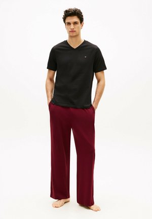Hombre de pie descalzo, vestido con una camiseta negra de cuello en V y pantalones ancho de color vino tinto, con las manos en los bolsillos frente a un fondo blanco liso.