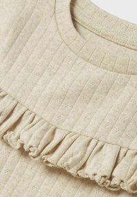 Lichtbeige top met een ribbelstructuur en kleine stippenpatronen, en een gerimpelde zoom langs de halslijn. Zachte katoenen stof.