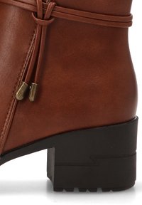 Botín de cuero marrón con acabado mate, detalle de lazada decorativa y un tacón negro grueso con suela texturizada.