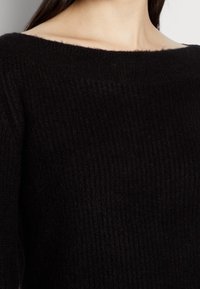 Czarny prążkowany sweter dzianinowy z łódkowym dekoltem, charakteryzujący się miękką teksturą i bez widocznych ozdób czy elementów metalowych. Zbliżenie na tkaninę.