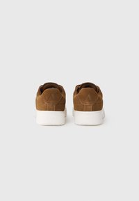 Brown suede sneakers featuring a low-top design, white rubber soles, and embossed branding at the heel. Adjustable lacing system.