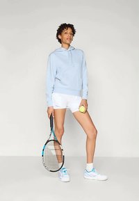 Žena u svijetloplavoj dukserici i bijelim šortsevima drži tenisku rekete i lopticu, stojeći pred jednobojnom sivom pozadinom.