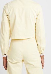 Vista trasera de una persona vestida con una chaqueta corta de color amarillo pálido y pantalones de talle alto a juego, de pie contra un fondo claro y liso.