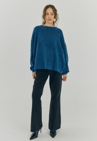 Modra oversized volnena jopica z okroglim izrezom in spušenimi rameni, v kombinaciji z visokimi temnimi kavbojkami in črnimi špicastimi petkami.