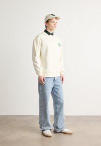 Felpa crema con grafica verde, camicia scura a colletto sotto, jeans di denim blu chiaro, sneakers beige e cappellino da baseball crema.