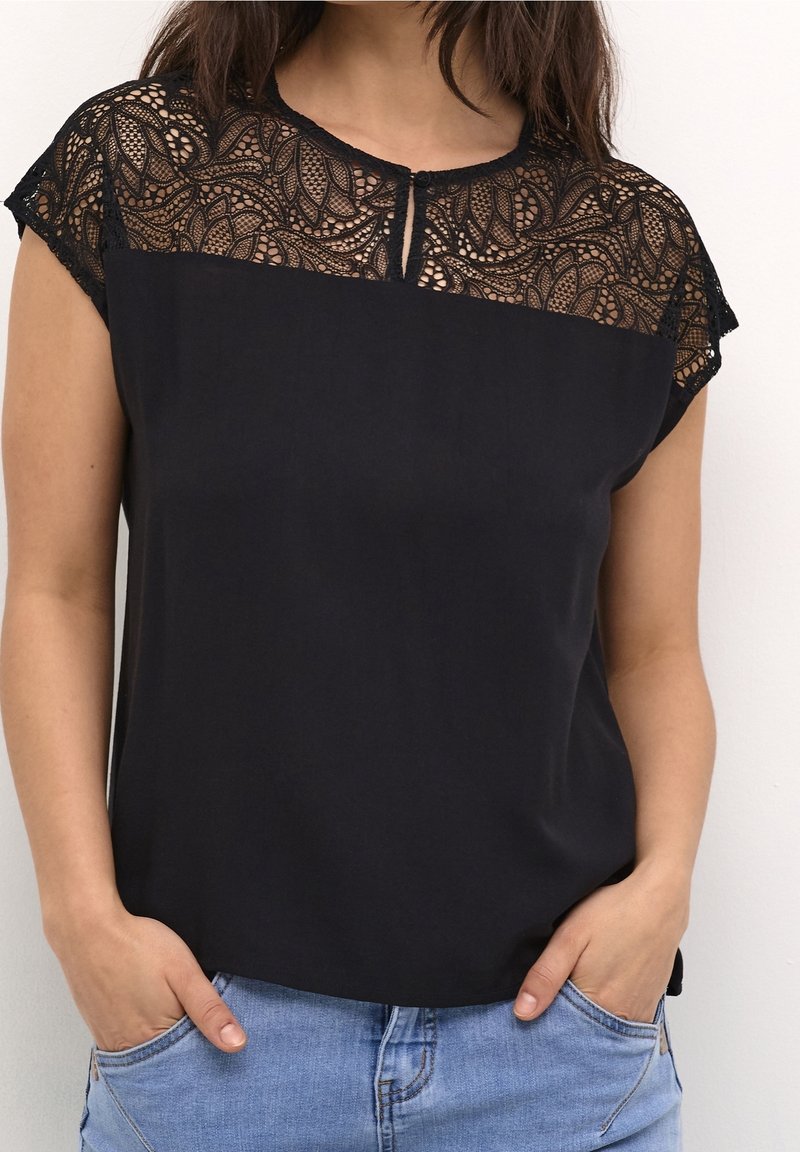 Donna che indossa un top nero a maniche corte con dettagli in pizzo intrecciati sulla parte superiore del petto e delle spalle, abbinato a jeans azzurri chiari.