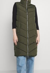 Oliwkowa pikowana kamizelka z wysokim kołnierzem, srebrnym zamkiem błyskawicznym i wzorem w jodełkę; posiada boczne kieszenie i sięga do kolan.