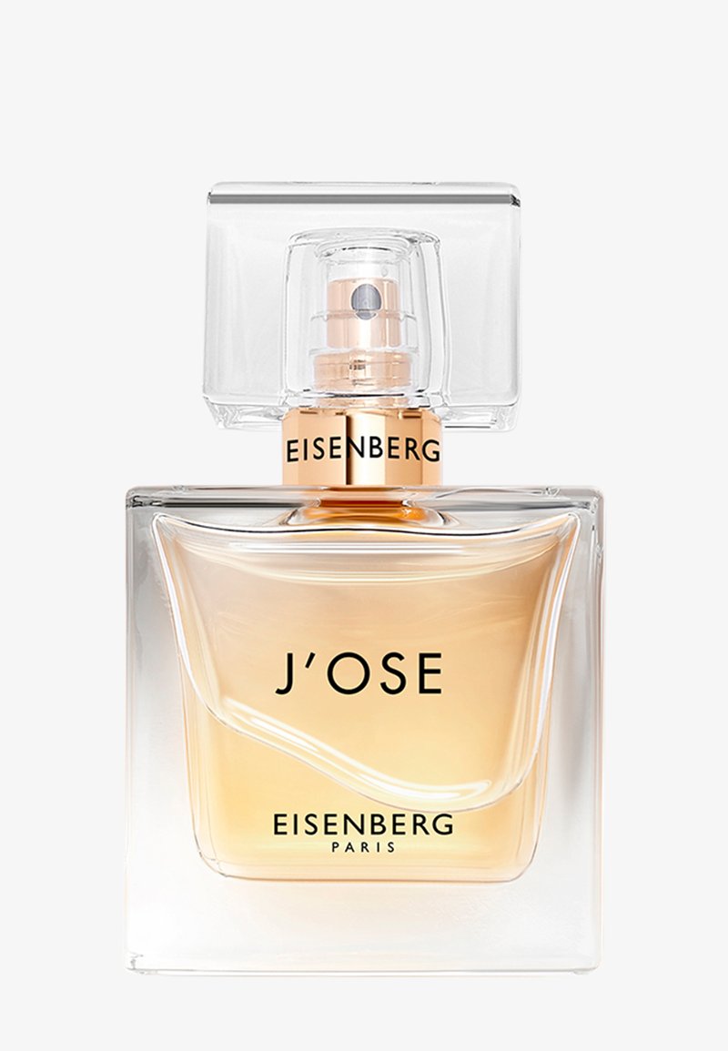 Bottiglia di profumo in vetro con un tappo quadrato trasparente. Il liquido è di un colore ambra chiaro, caratterizzato da un etichetta con testo nero audace e un accento dorato.