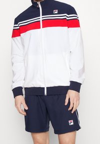 White athletic jacket with navy and red stripes, zippered front, and Fila logo. Paired with navy shorts featuring white side piping and logo.