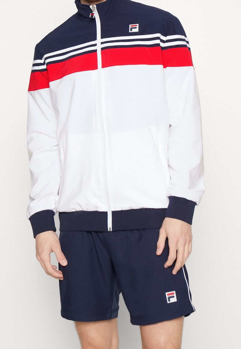 White athletic jacket with navy and red stripes, zippered front, and Fila logo. Paired with navy shorts featuring white side piping and logo.
