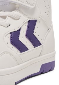 Weißer Sportschuh mit perforierter Textur, ausgestattet mit lila Zickzack-Akzenten und einer stabilen Sohle. Klettverschluss und Schnürsenkel sind sichtbar.