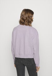 Felpa cropped di colore viola chiaro con bordo e polsini a coste. Presenta maniche ampie e un piccolo fiocco decorativo sulla parte posteriore del collo.