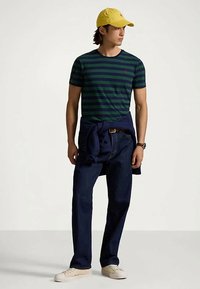 Άνδρας με κίτρινο καπέλο, ναυτικό και πράσινο ριγέ μπλουζάκι, σκούρα τζιν, λευκά sneakers και ναυτικό φούτερ δεμένο στη μέση.