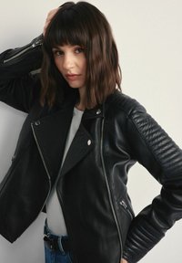 Czarna skórzana motocyklowa kurtka z żebrowanymi akcentami na ramionach, asymetrycznym zamkiem błyskawicznym i gładką powierzchnią, połączona z jasną bluzką i dżinsami.