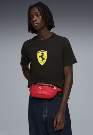 Camiseta negra con logo de Ferrari amarillo. Riñonera roja con un compartimento con cremallera, con un pequeño logo, llevada sobre vaqueros oscuros.