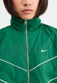 Zaļš Nike jaka ar augstu apkakli, priekšu ar rāvējslēdzēju, baltām apdares detaļām un logo. Izgatavota no viegla, teksturēta auduma.