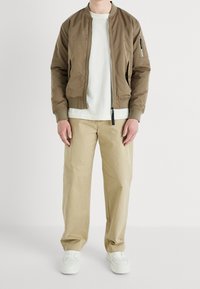 Man in een tan bomberjack over een witte t-shirt, losse beige broek en witte sneakers, staande tegen een effen witte achtergrond.