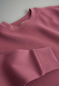 Tuva skatup augšdaļas vaļējs, ar redzamu šuvumu un salocītu piedurkni uz pelēka fona.