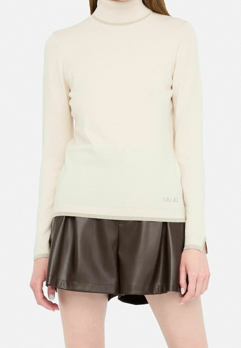 Femme portant un pull à col roulé crème associé à un short plissé en cuir marron foncé sur fond blanc.