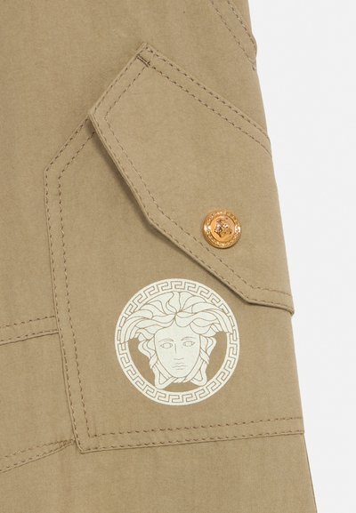 Tela beige con solapa de bolsillo cosida, botón dorado y un emblema blanco con la cabeza de Medusa encerrado en un círculo de clave griega.