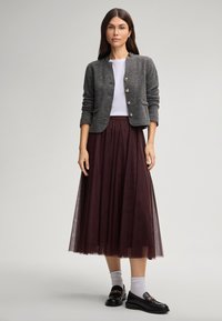 Woman wearing a grey buttoned cardigan, white top, burgundy midi tulle skirt, white socks, and black loafers standing against a plain background.