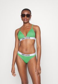 Zelené bikiny s trojúhelníkovým vrškem a odpovídajícími spodními díly, které mají bílý pás s logem "TOMMY JEANS". Hladký materiál, minimalistický design.