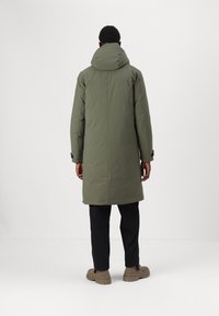 Langer olivgrüner Daunenmantel mit Kapuze, gerader Schnitt und minimalistisches Design. Getragen über dunklen Hosen und hellen Schuhen.