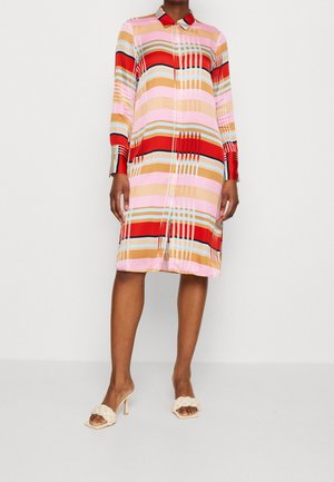 Femme portant une robe boutonnée à hauteur de genou avec des rayures multicolores horizontales et verticales, et des sandales à talons beiges avec des brides tressées.