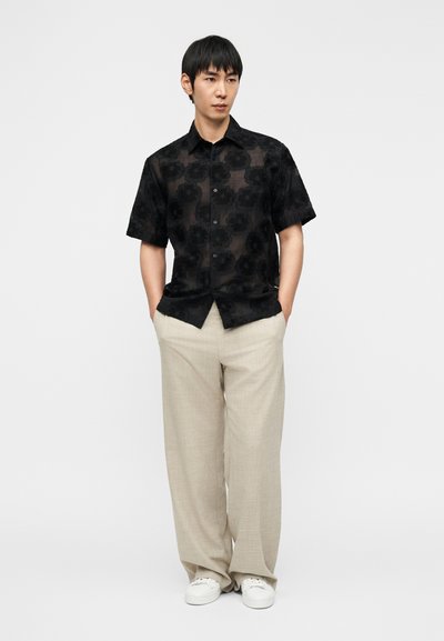 Πουκάμισο με κοντά μανίκια, μαύρο με λουλουδένιο μοτίβο και κουμπιά, συνδυασμένο με ανοιχτό μπεζ παντελόνι με φαρδιά μπατζάκια και λευκά αθλητικά παπούτσια.