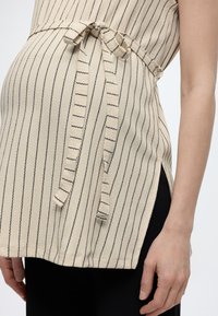 Beige ermeløs topp med tynne, svarte, vertikale striper, knytt belte i midjen og sidespalt over svarte bukser på en persons overkropp og arm.