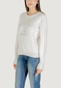 Witte trui met het "GUESS" logo in zilveren rhinestones, lange mouwen en geribbelde zoom, gecombineerd met blauwe denim jeans. Gladde textuur.