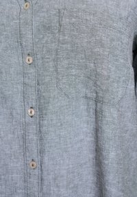 Leinenhemd in Hellgrau mit einer subtilen Textur, ausgestattet mit einer Tasche und natürlichen, holzähnlichen Knöpfen entlang der Vorderseite.