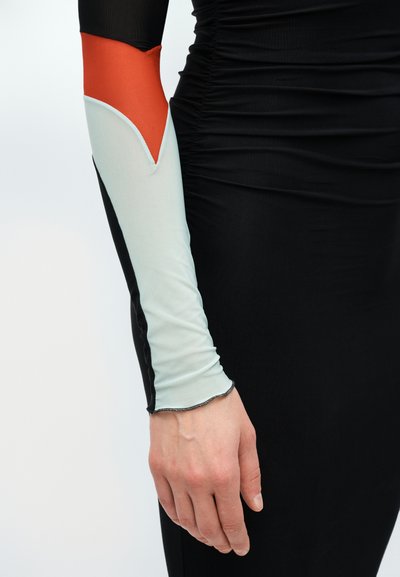 Kar hosszú ujjú ruhadarabban fekete, fehér és narancssárga színblokkokkal, mellett ráncolt fekete anyag egy ruhán vagy felsőn.