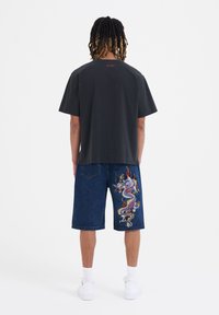 Black cotton t-shirt and blue denim shorts. Shorts feature a colorful dragon embroidery on the right leg. Model wears white sneakers.