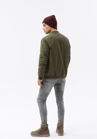 Giacca bomber verde oliva con polsini a coste, jeans skinny grigi sbiaditi e stivali marroni con lacci verde brillante, modello visto da dietro.