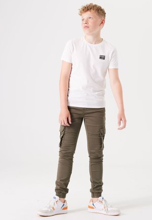 Weißes T-Shirt mit kurzen Armen und einem kleinen schwarzen Etikett, kombiniert mit olivgrünen Cargo-Hosen mit Seitentaschen und Knöchelbündchen. Weiße Sneaker mit orangefarbenen Akzenten.