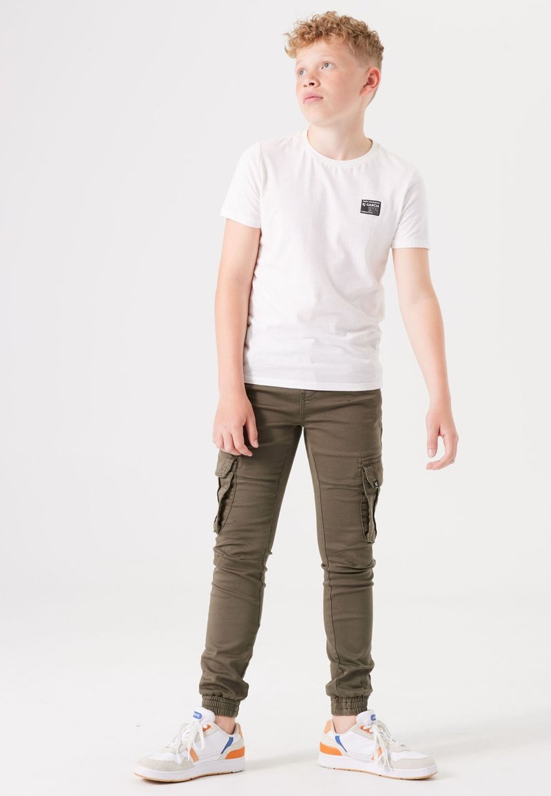 Weißes T-Shirt mit kurzen Armen und einem kleinen schwarzen Etikett, kombiniert mit olivgrünen Cargo-Hosen mit Seitentaschen und Knöchelbündchen. Weiße Sneaker mit orangefarbenen Akzenten.
