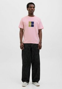 Lyserød t-shirt med korte ærmer og et firkantet grafik i grøn, blå og hvid tekst, parret med mørke, brede bukser og hvide sneakers.