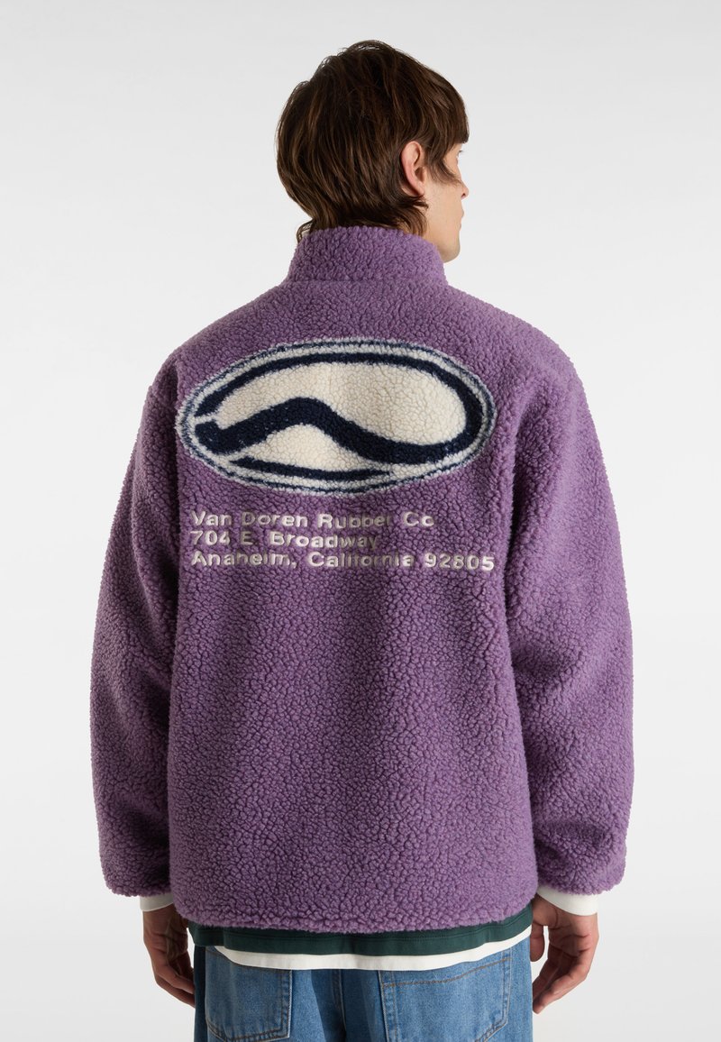 Lila Fleecejacke mit hohem Kragen. Auf der Rückseite prangt ein großes Wellenlogo in Weiß und Marineblau. Unter dem Logo steht ein Text.