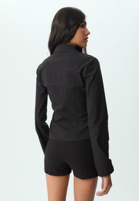 Čierny blejzer s jemným pruhovaním a dlhými rukávmi, slim-fit strihom a špicatým golierom, skombinovaný s čiernymi šortkami. Hladká textúra, elegantná silueta.