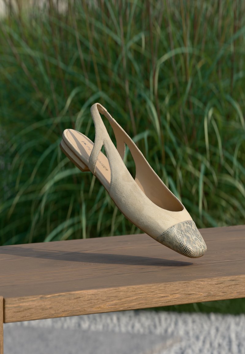 Μπεζ επίπεδο παπούτσι slingback με υφή και αιχμηρή μύτη, που αιωρείται πάνω από ξύλινο παγκάκι με πράσινα φυτά στο φόντο.