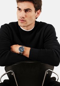 Montre argentée avec cadran bleu au poignet, présentant des détails de chronographe. Pull texturé sombre, assis sur une chaise noire avec des accents métalliques.