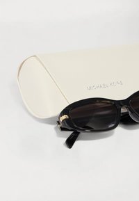 Occhiali da sole neri con lenti scure e aste con inserti dorati, posizionati accanto a una custodia bianca texturizzata con la scritta "MICHAEL KORS" in rilievo.