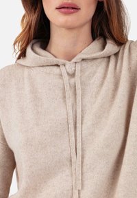 Helles beiges Kapuzen-Sweatshirt aus weichem Strickstoff. Verfügt über eine Kapuze mit Kordelzug und eine lockere Passform mit gerippten Details entlang des Halsausschnitts und Saums.