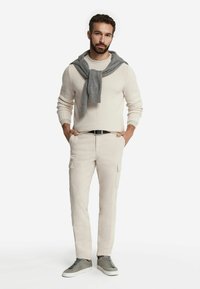 Lichtbeige cargobroek met zijzakken, gecombineerd met een gestructureerde off-white trui en een grijze sweatshirt over de schouders, afgewerkt met grijze sneakers.
