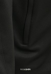 Close-up van zwarte stof met een verticale naad en een klein wit "BEECOOL"-logo vlak bij het onderste midden.