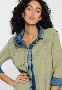 Green and blue denim shirt with a prominent collar, button closure, and chest pocket. Features contrasting fabric textures and rolled sleeves.