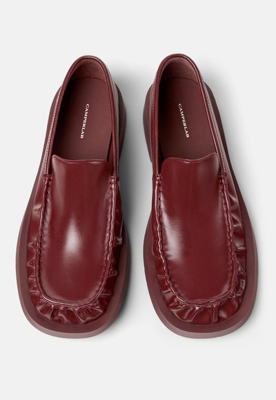 Par de mocasines de cuero rojo oscuro brillante sin cordones, con detalle de pliegues en el borde y suela gruesa, vistos desde arriba sobre un fondo blanco.